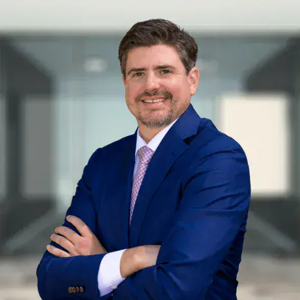 Hombre con traje azul y corbata rosa claro de pie con los brazos cruzados, sonriendo, delante de un fondo borroso de oficina moderna.