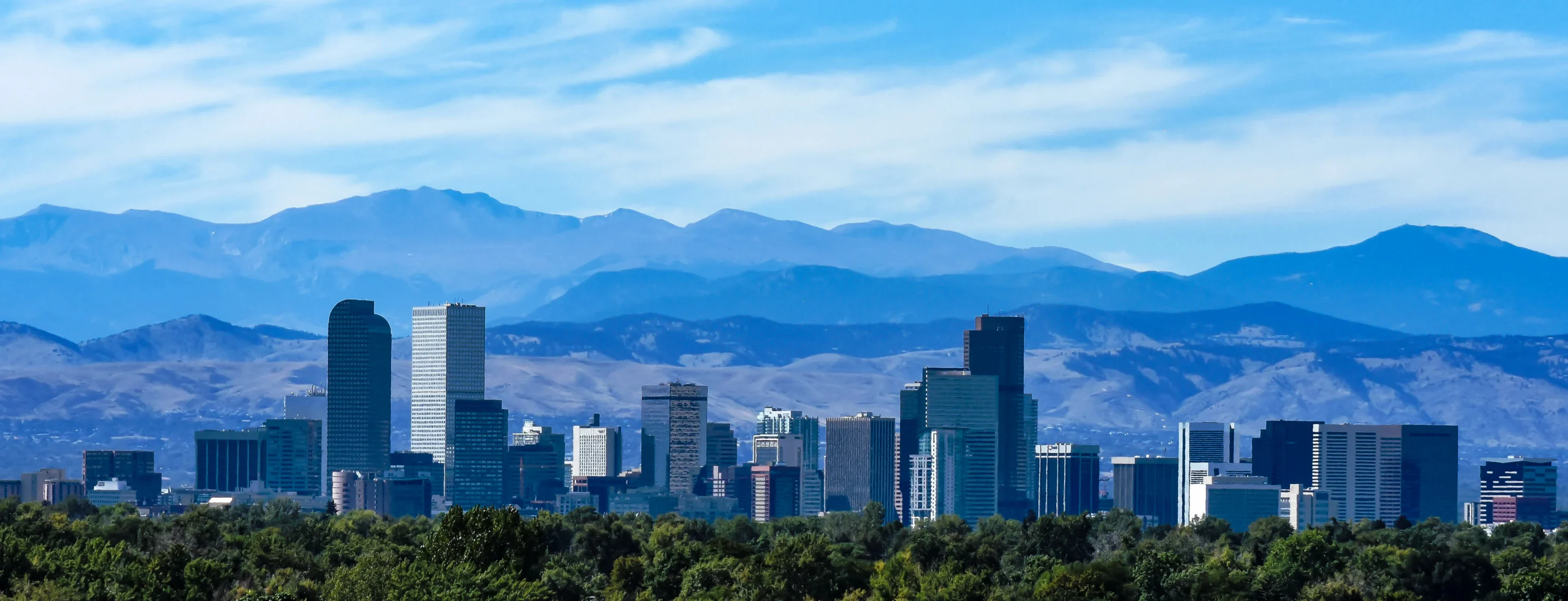 A city skyline featuring tall buildings with mountains and a blue sky in the background, and trees in the foreground.