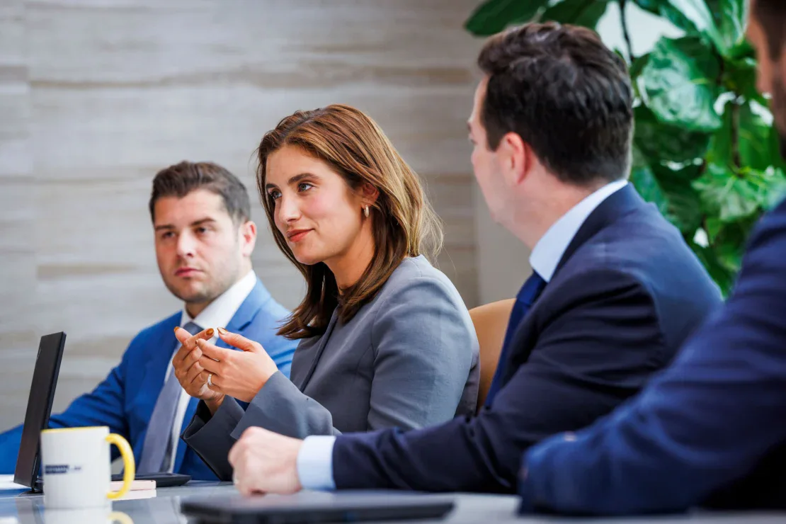 Three people in business attire sit at a conference table with laptops, engaged in a meeting or discussion.