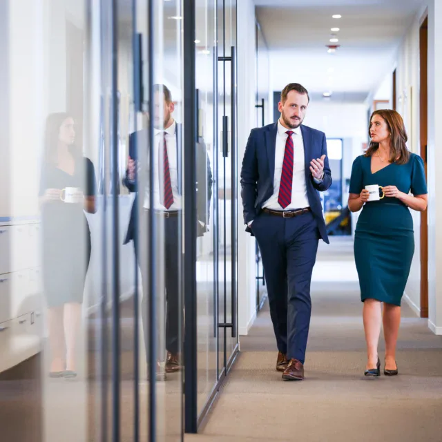 Two professionally dressed people walk and talk in an office hallway, holding coffee cups and appearing engaged in conversation.