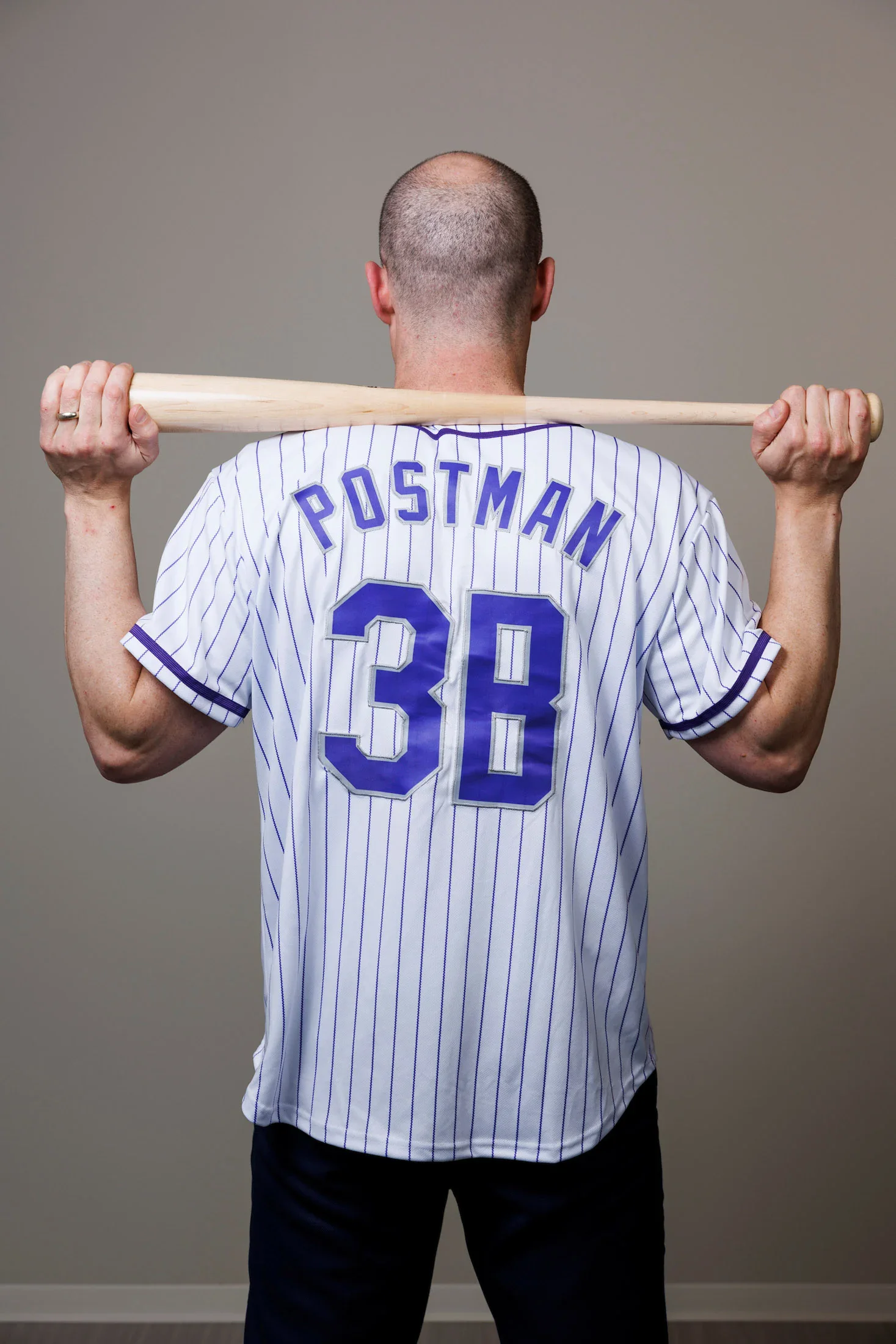 Una persona está de espaldas a la cámara, sosteniendo un bate de béisbol detrás de los hombros. Lleva una camiseta blanca a rayas con las palabras "POSTMAN" y "3B" en la espalda.