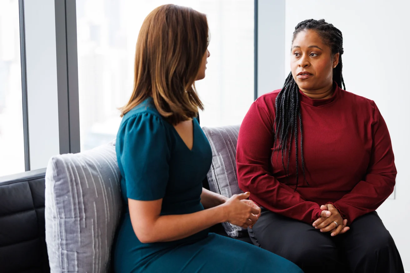 Two women sit on a couch in conversation; one wears a teal dress and the other a red long-sleeve top. They appear engaged in a serious discussion.