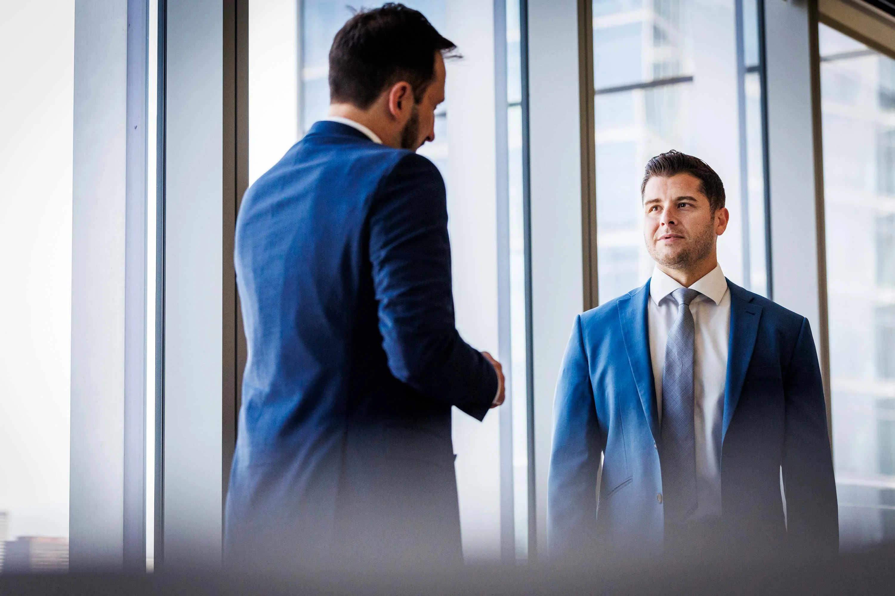 Two men in business suits have a conversation in a modern office with large windows and natural light.