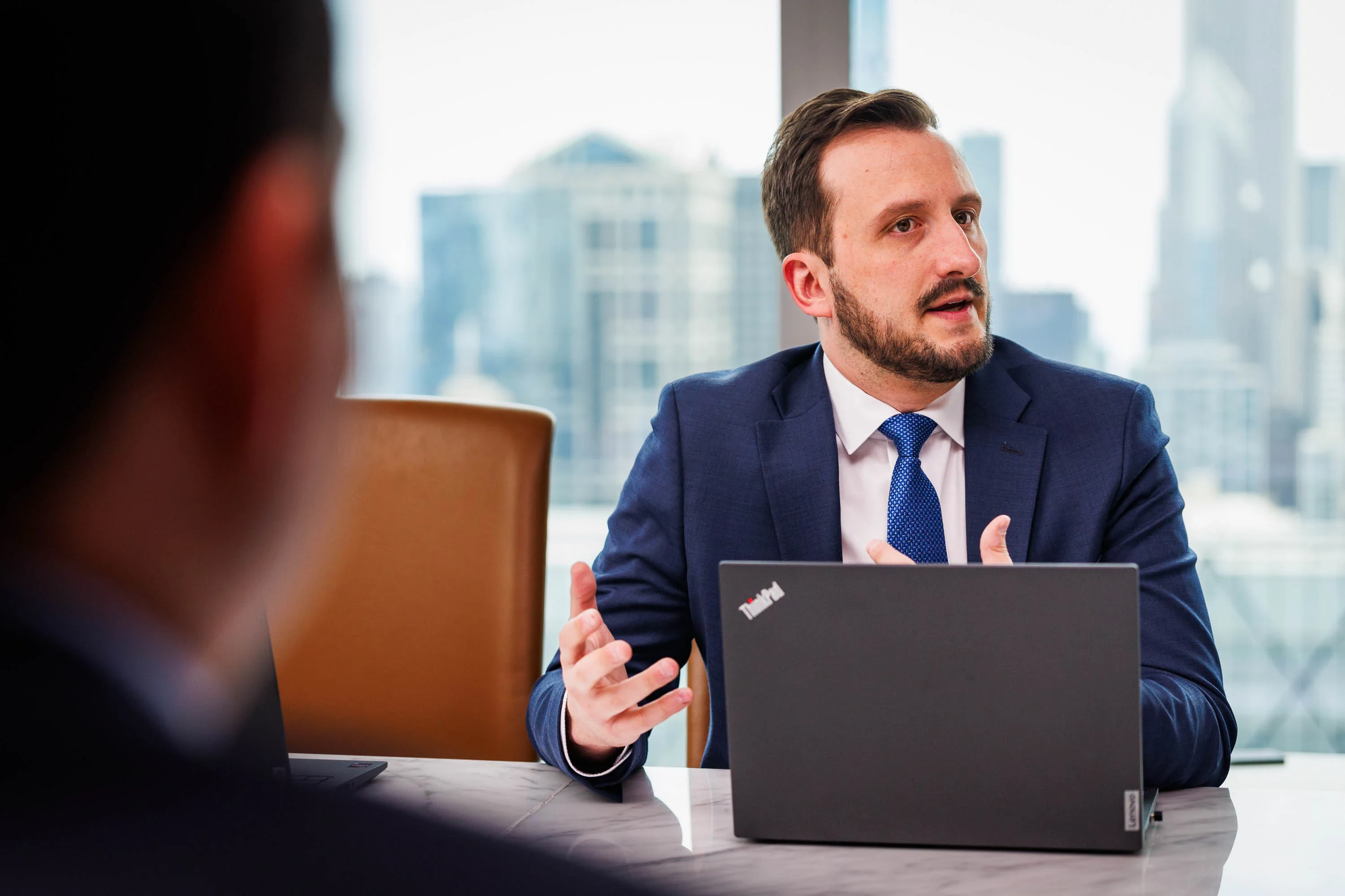 A man in a suit sits at a conference table with a laptop, gesturing as he speaks during a business meeting.