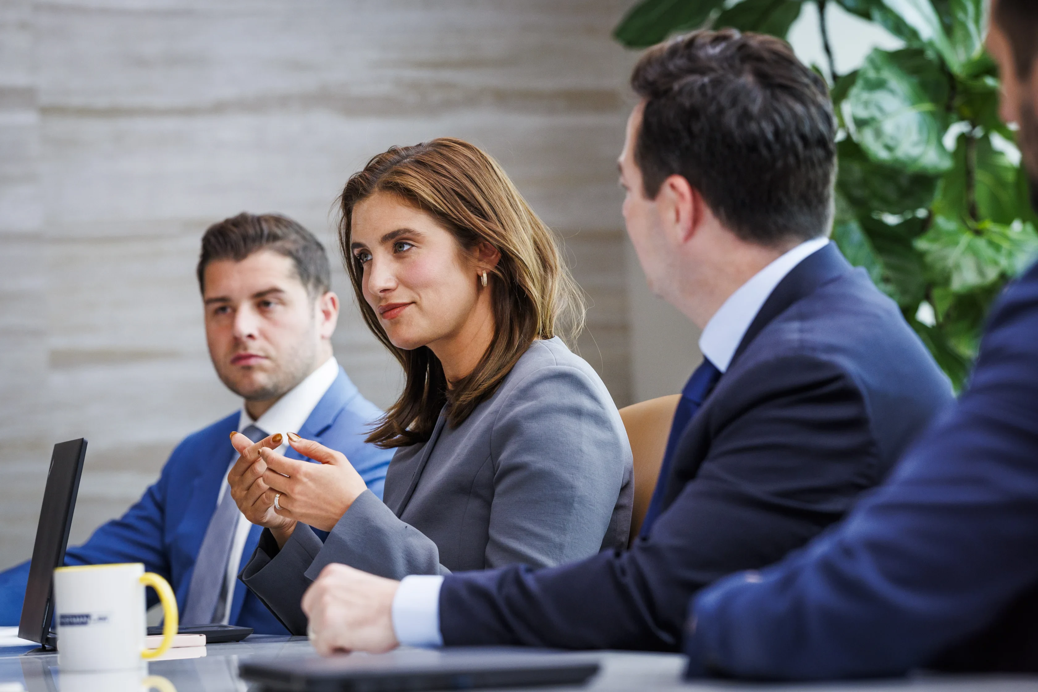 Three people in business attire sit at a conference table with laptops, engaged in a discussion during a meeting.