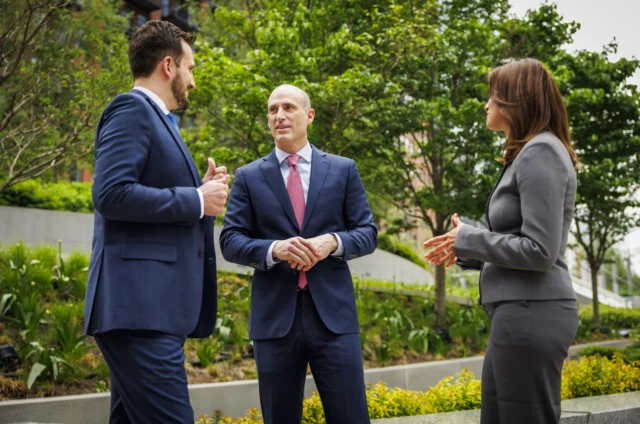 Three people in business attire stand outside having a discussion in front of greenery and modern buildings.