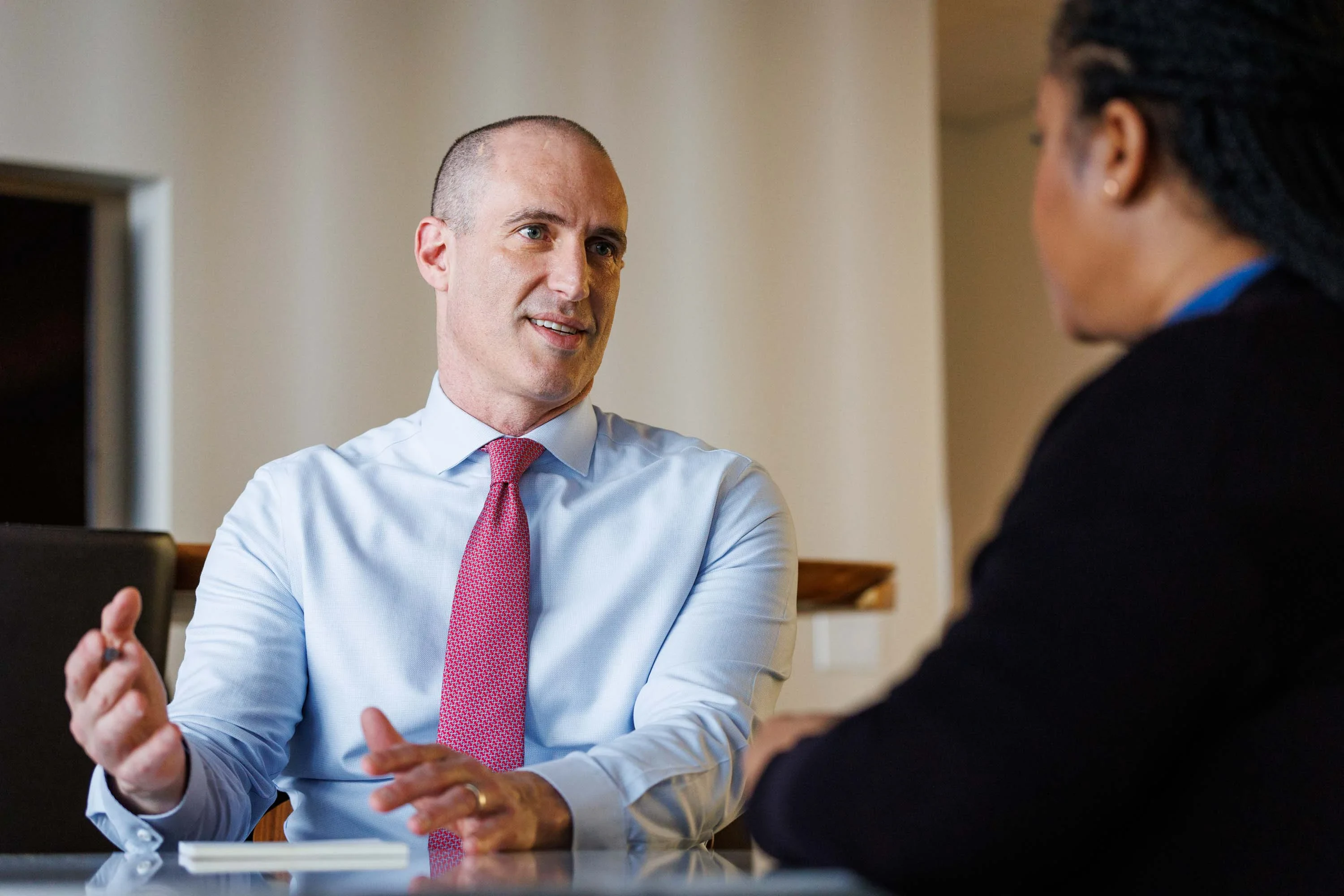 A man in a light blue shirt and pink tie speaks to a woman during a conversation at a table in an office setting.