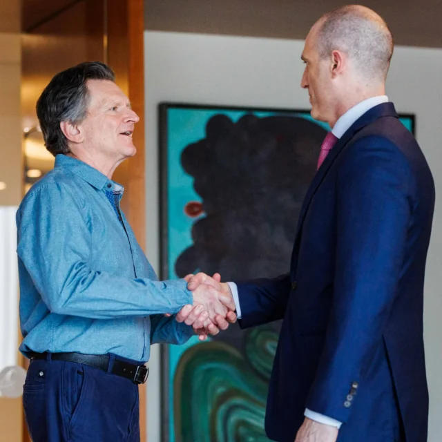 Two men shake hands indoors, one wearing a blue shirt and jeans, the other in a navy suit; abstract artwork and a plant are visible in the background.