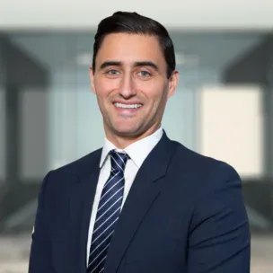 Hombre con traje azul marino y corbata a rayas de pie en una oficina, sonriendo a la cámara.