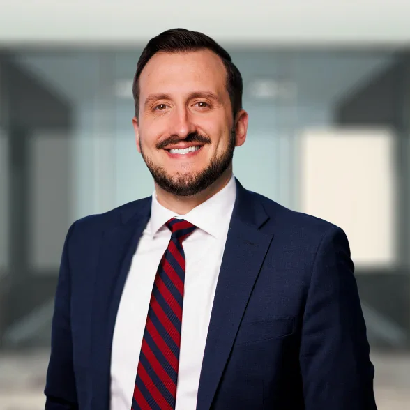 A man in a navy suit, white shirt, and red striped tie smiles while standing in an office setting with glass walls.
