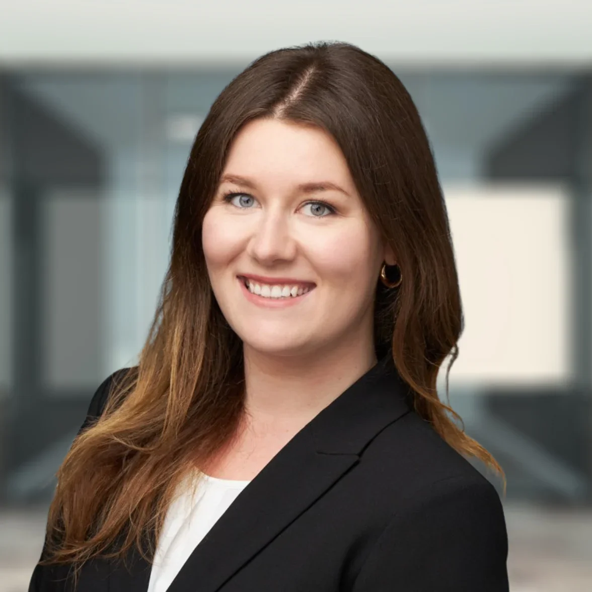 Woman with long brown hair wearing a black blazer and white top, smiling, standing in a modern office setting.