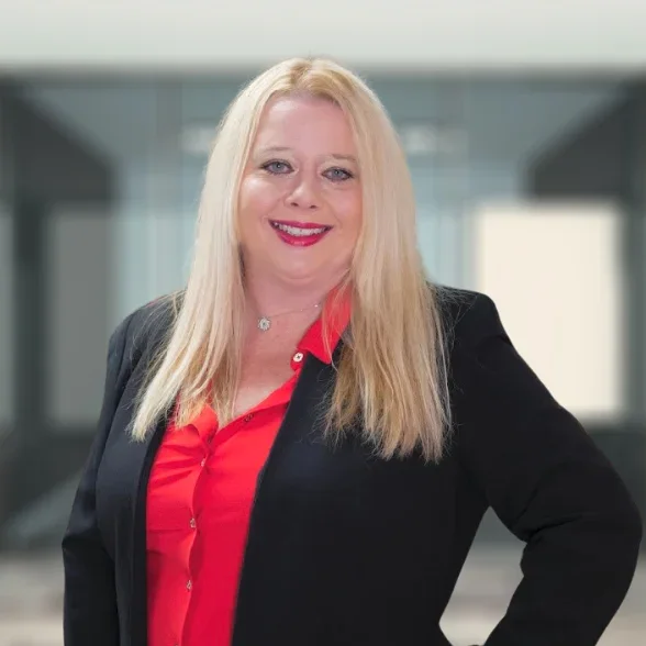 A woman with long blonde hair, wearing a black blazer over a red blouse, stands smiling in a modern office hallway.