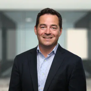 Un hombre con traje azul marino y camisa azul claro sonríe, de pie delante de un fondo de oficina difuminado.