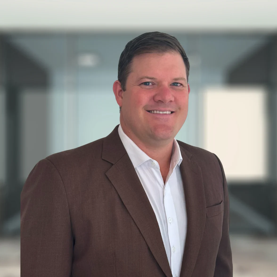A man in a brown suit jacket and white shirt stands in a modern office setting, smiling at the camera.