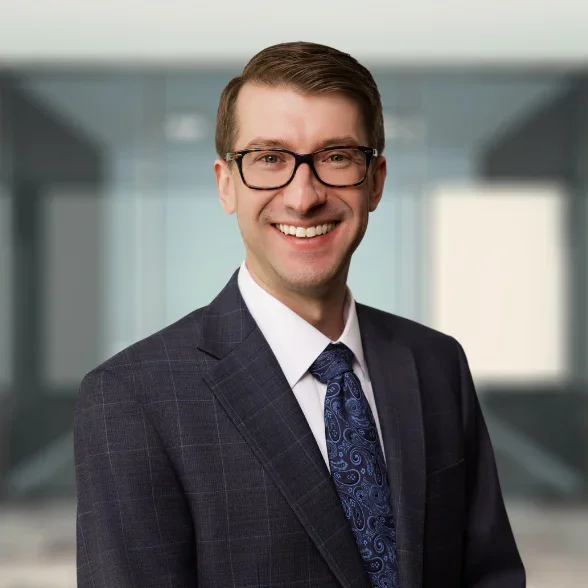 A man in a suit and tie, wearing glasses, smiles while standing in front of a blurred office background.