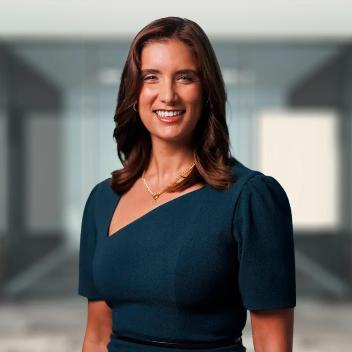 A woman with brown hair in a dark teal dress stands smiling in a modern office setting.