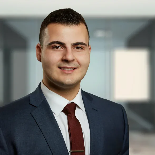 A man in a navy blue suit, white shirt, and burgundy tie is smiling in a modern office setting with blurred glass walls in the background.