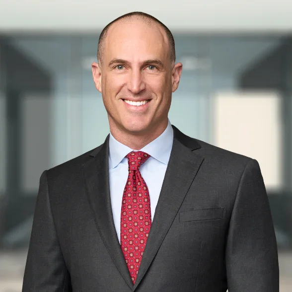 A man in a dark suit, light blue shirt, and red patterned tie, smiling, stands in front of a blurred office background.