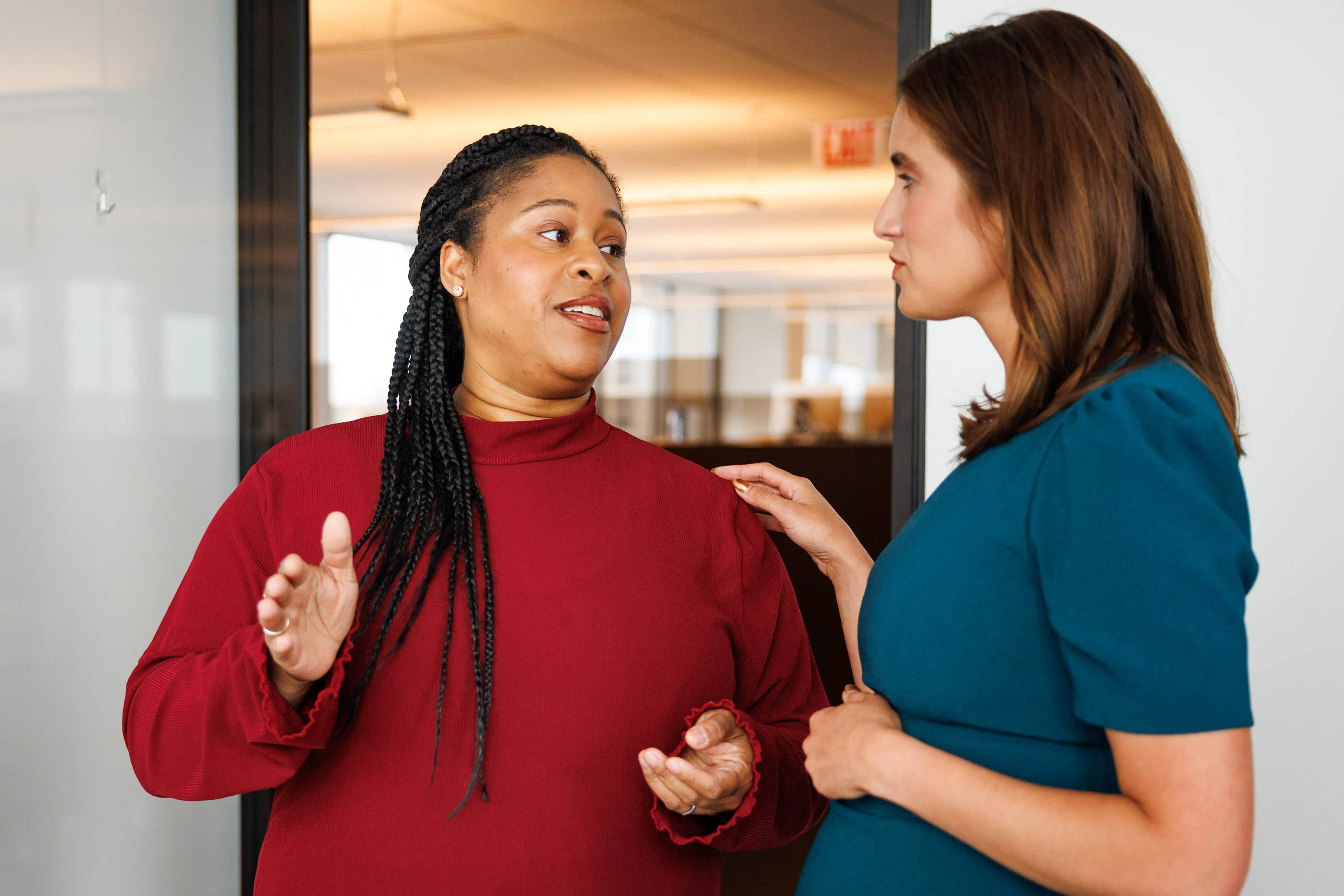 Two women stand indoors having a serious conversation; one gestures with her hands while the other rests her hand on her shoulder.