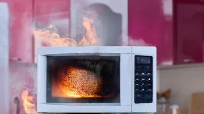 A microwave oven on a countertop is on fire, with visible flames and smoke coming from inside and around the appliance in a kitchen setting.