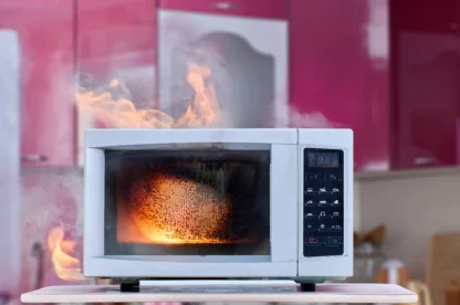 A microwave oven on a countertop is on fire, with visible flames and smoke coming from inside and around the appliance in a kitchen setting.