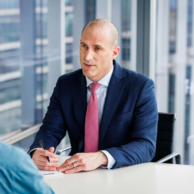 A man in a suit and tie sits at a desk with a pen and paper, speaking to another person in a modern office setting with large windows.
