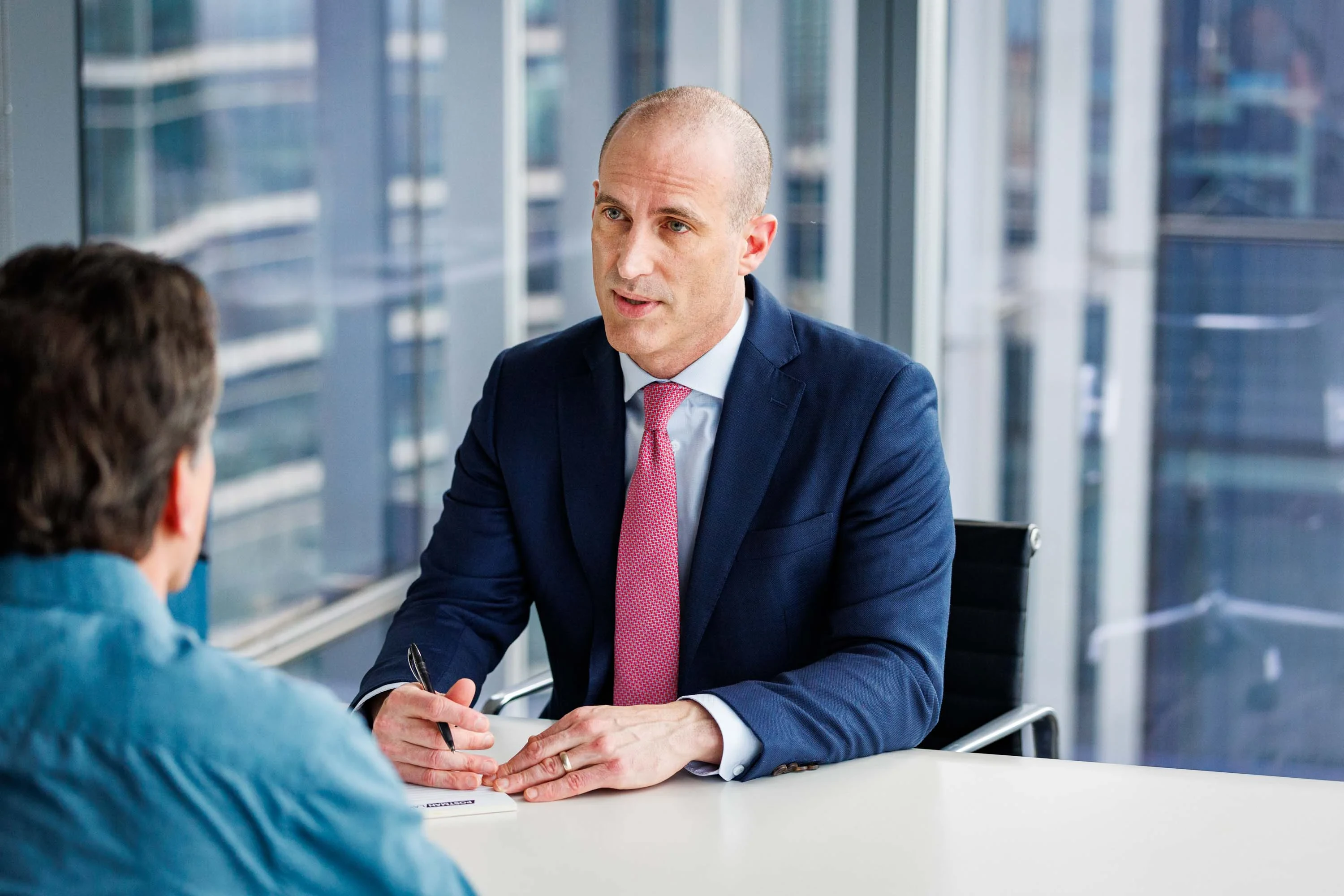 A man in a suit and tie sits at a desk with a pen and paper, speaking to another person in a modern office setting with large windows.