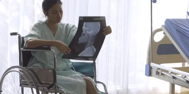 A person in a hospital gown sits in a wheelchair, examining an X-ray image of a skull next to a hospital bed.