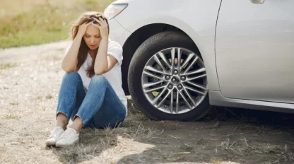 A woman sits on the ground next to a car with an open hood, holding her head in her hands and appearing distressed.