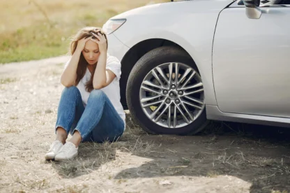 A woman sits on the ground next to a car with an open hood, holding her head in her hands and appearing distressed.