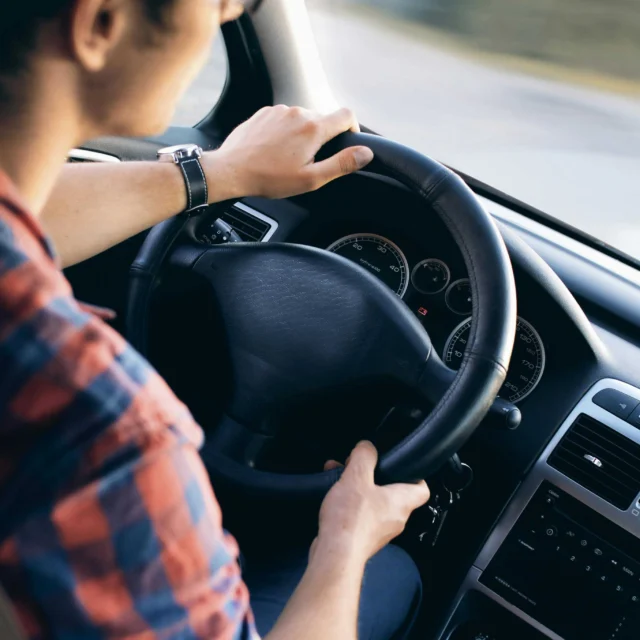 Person driving a car, hands on the steering wheel, wearing a plaid shirt; the dashboard and interior controls are visible.