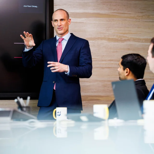 A man in a suit presents medical imagery on a screen to three colleagues seated at a conference table with laptops and coffee mugs.