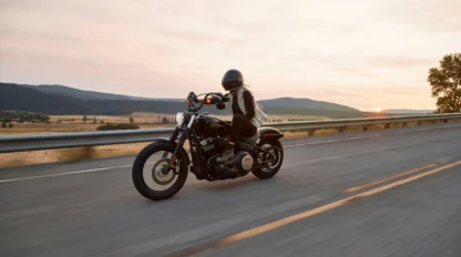 A person in a helmet rides a black motorcycle on an open highway at sunset with hills and fields in the background.