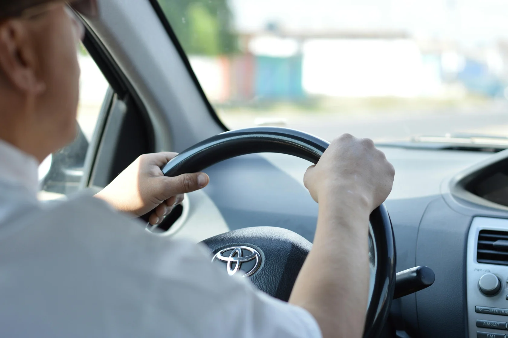 Person driving a Toyota car, hands on the steering wheel, view from the passenger seat with dashboard and part of the windshield visible.