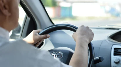 Person driving a Toyota car, hands on the steering wheel, view from the passenger seat with dashboard and part of the windshield visible.
