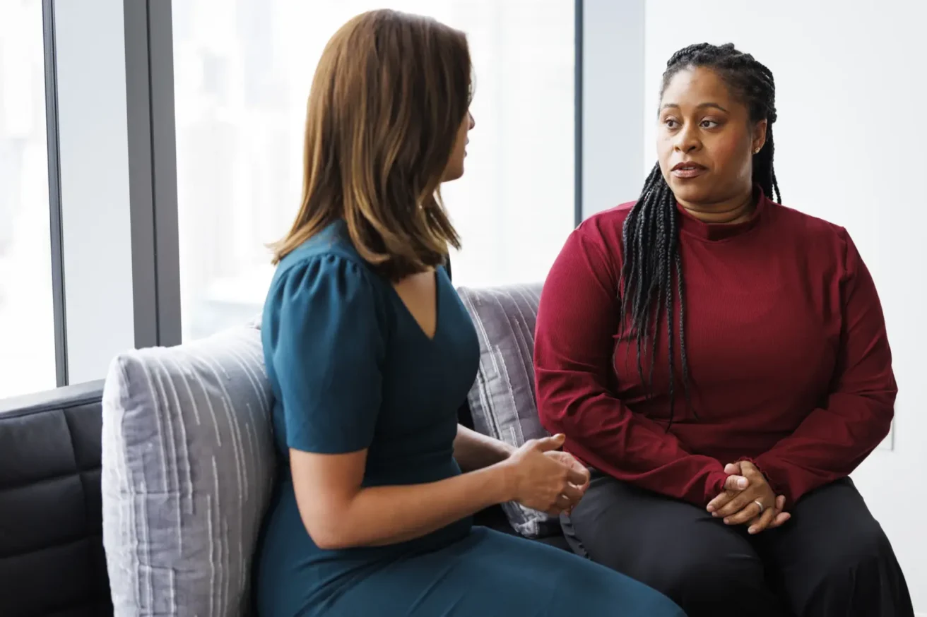 Two women sit on a couch having a serious conversation near large windows in a bright room.