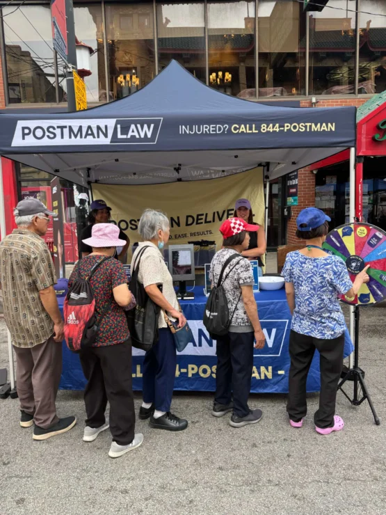A group of elderly people stand in line at a Postman Law booth on a city street, with a staff member assisting them and a prize wheel visible on the side.