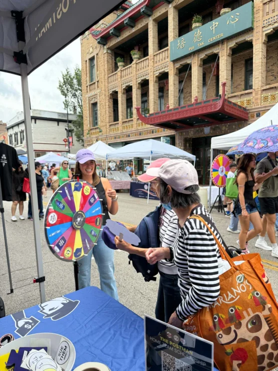 Two women stand at a booth on a city street, one spinning a colorful prize wheel while the other looks on. People and vendor tents line the street in the background.